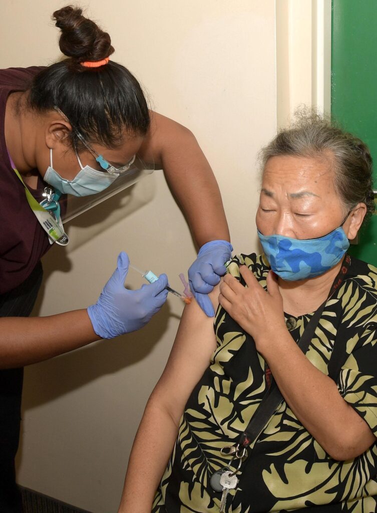 A senior sitting in an apartment hallway wearing a mask, receiving a vaccination from WoodGreen staff.