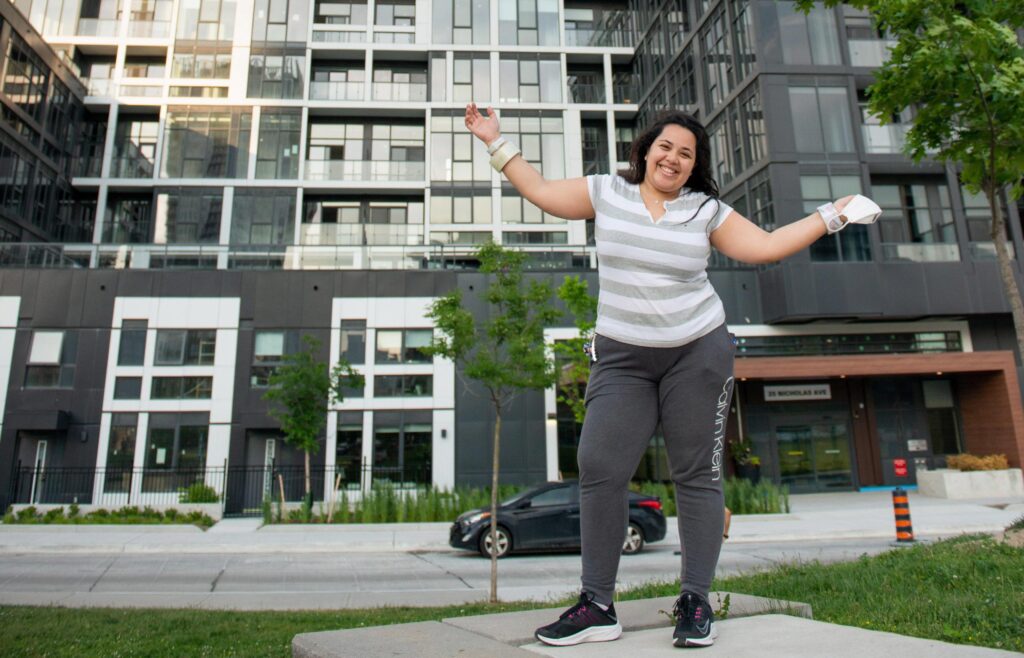 A woman posing outside an apartment building with her arms outstretched and proudly smiling.