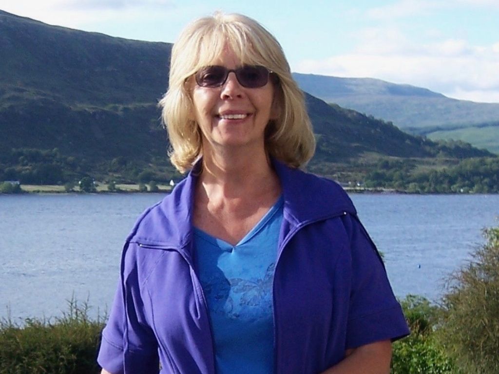 A woman in a blue shirt and a purple cardigan stands in front of a lake in Scotland