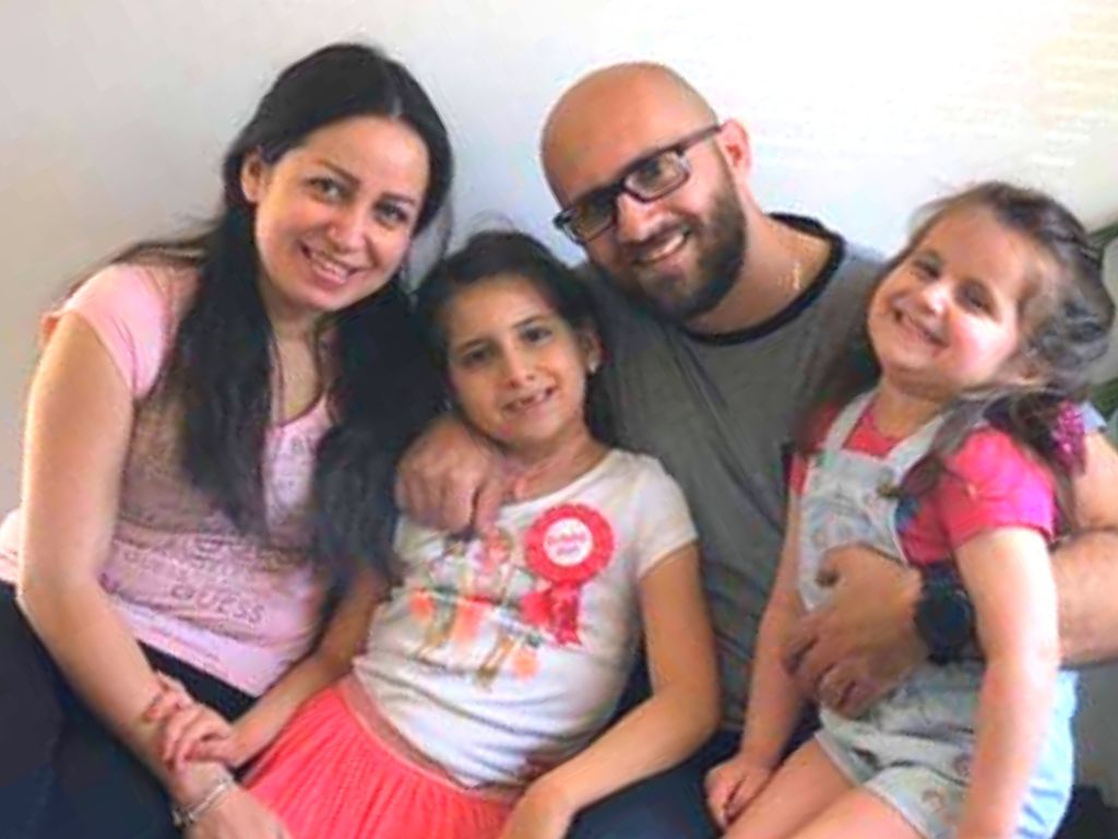 A family with a mother, father, and two young daughters sits smiling on a couch. 