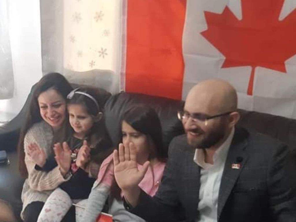 A family with a mother, father and two young daughters raise their hands to swear an oath in front of a Canadian flag. 