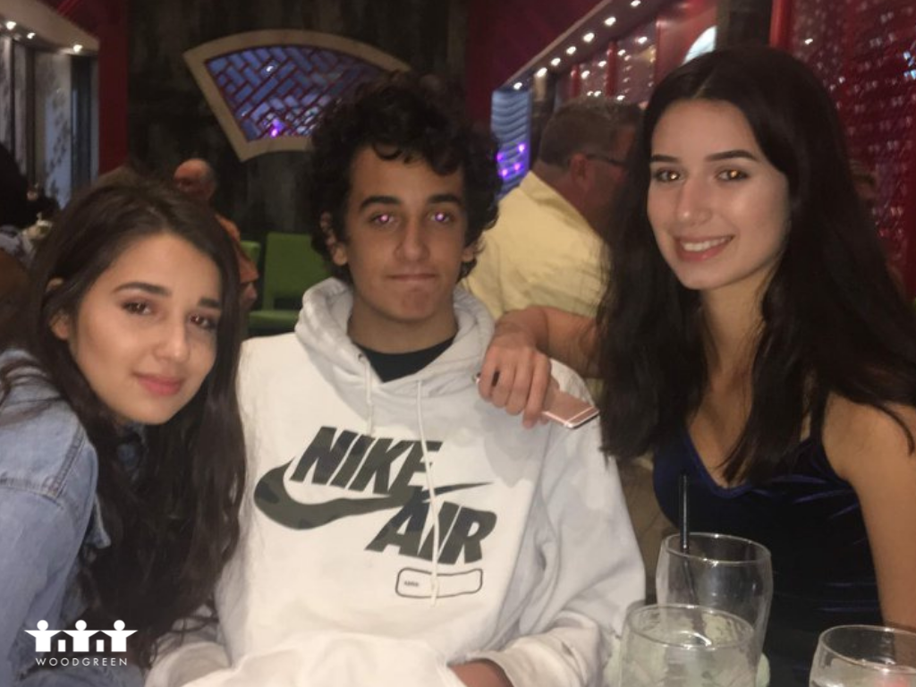 a teenage girl with long dark hair, a teenage boy with short dark hair and a teenage girl with long dark hair sit with their arms around each other at a table