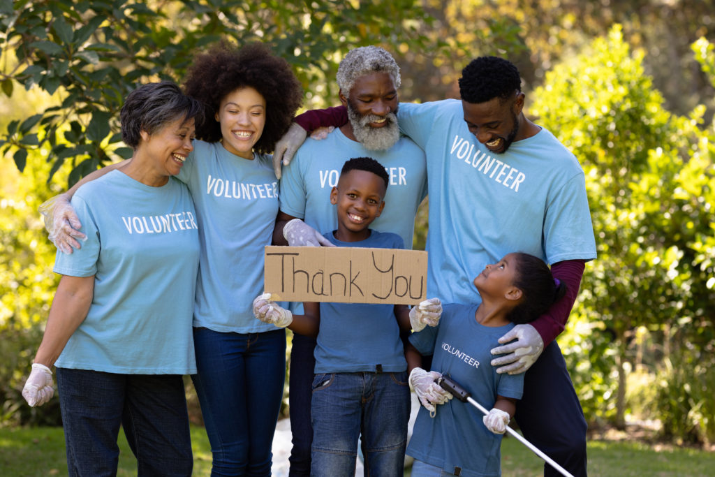 Multi-generation mixed race family spending time outside together, all wearing blue volunteer t shirts and protective gloves, collecting garbage, standing in a row, embracing each other, holding a volunteer sign, on a sunny day