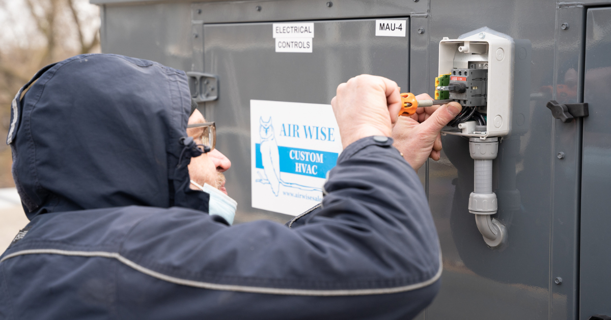 A man wearing a blue jacket uses a tool to repair an electrical unit. 