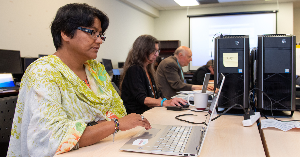 A woman with dark hair and skin, a woman with dark brown hair and light skin and an older white man with balding hair sit at computers and type.
