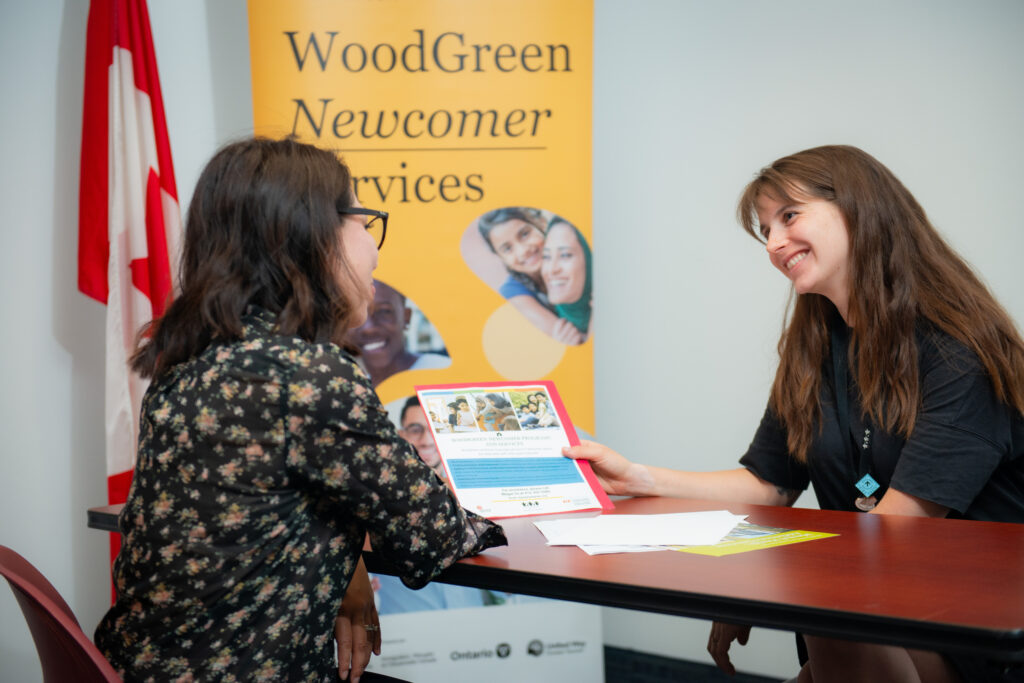 Woman showing a flyer to a client in front of a banner that reads Newcomer Services
