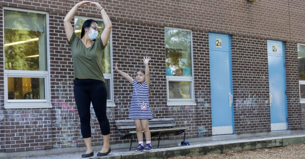 An Early Childhood Educator (ECE ) dances with a little girl in a blue and white striped dress on a bench outside in front of a brick wall. 