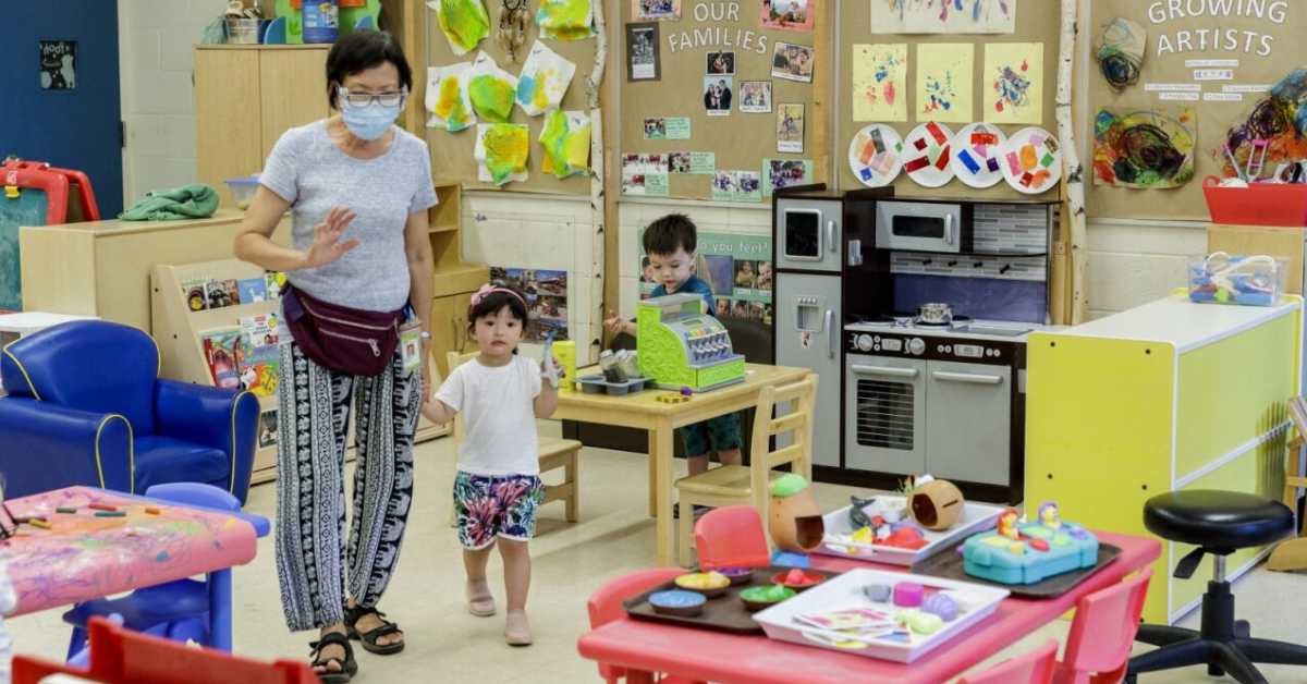 An Early Childhood Educator ECE wearing black and white pants, a grey shirt and a fanny pack holds a young child by the hand in a day care centre. 