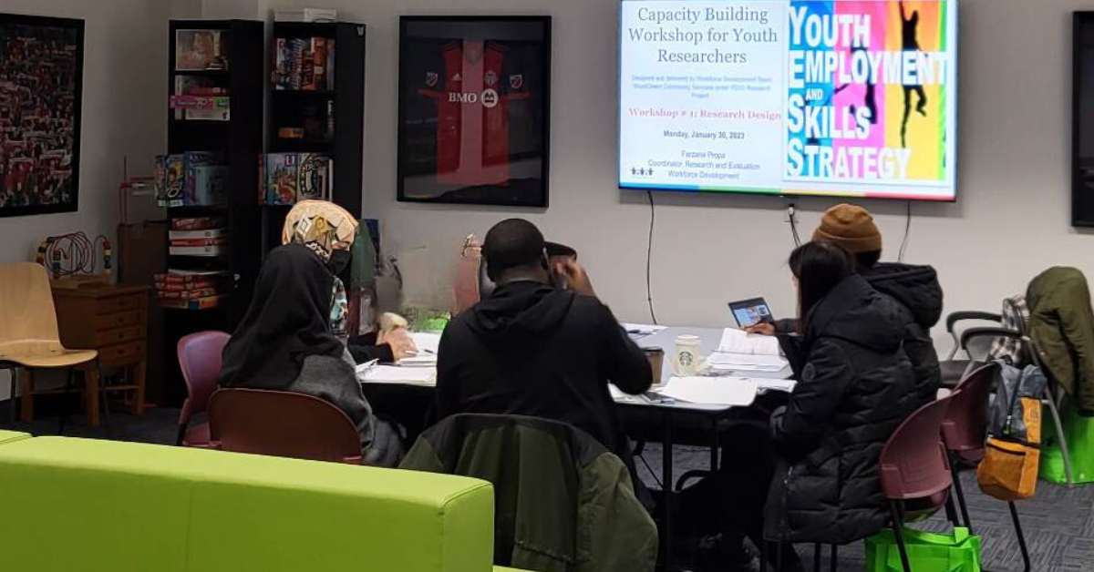 Five newcomer youth sit around a table in a conference room with their back to the camera while looking at a screen that says Youth Employment and Skills Strategy