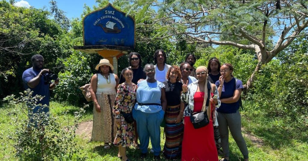 a group standing in a grassy patch near a tree, in front of a sign that says Coral Cavern Mangarpnani