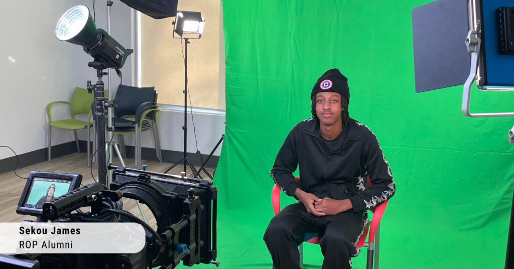 a young Black man smiling while being recorded on camera, sitting in front of a green screen