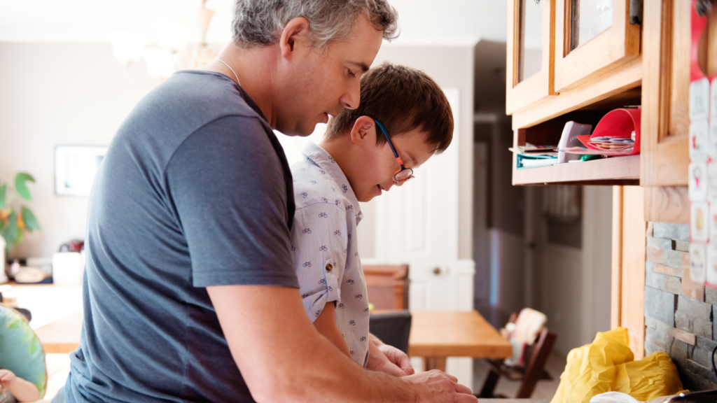 an older man in a blue shirt leans over a young boy in pyjamas and glasses in the kitchen.