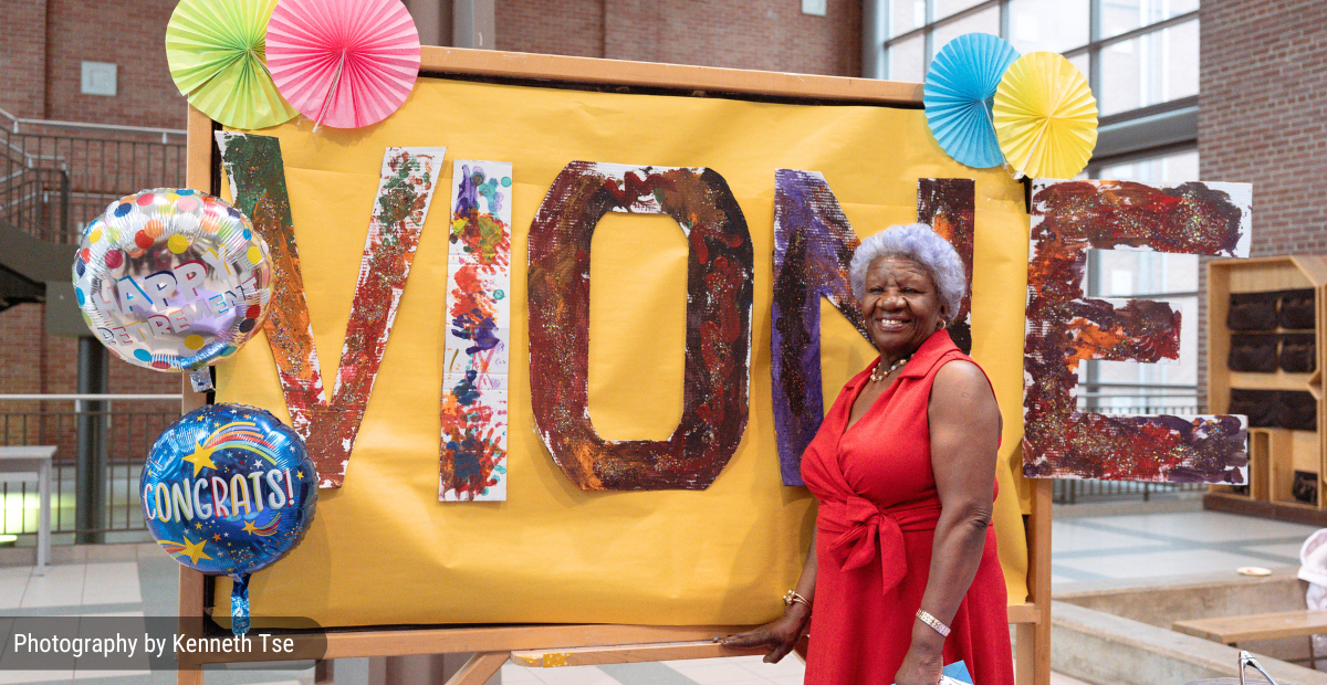 a woman with dark skin wearing a red dress stands in front of a sign painted by children that reads Vione.
