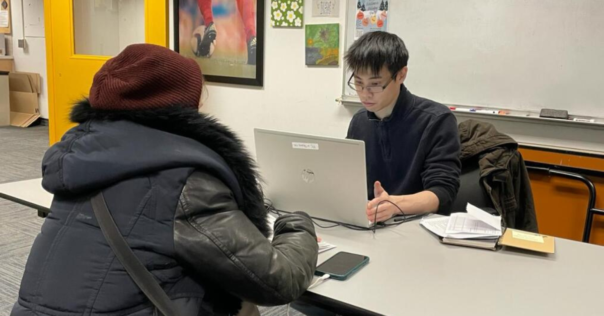 a woman in a black jacket sits at a desk facing a man on a laptop computer