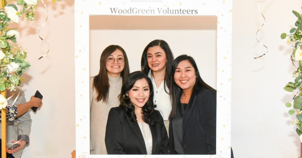 four women, all with long dark hair stand in a white cardboard frame reading WoodGreen Volunteers