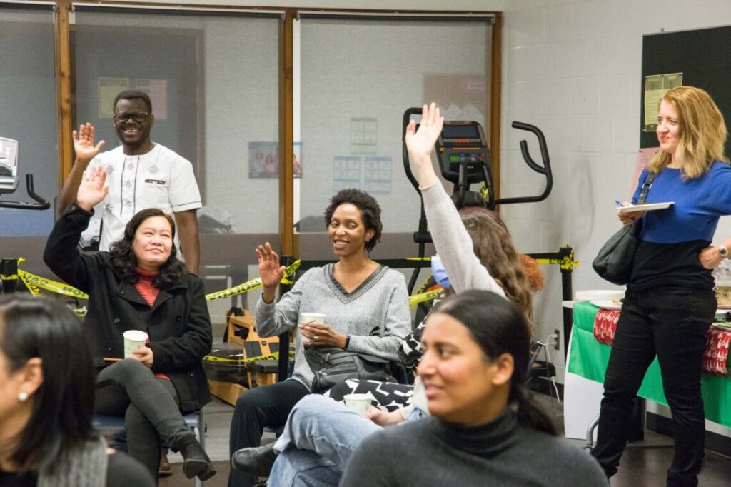 a man is standing in a conference room while 4 women are sitting with three of them raising their hands