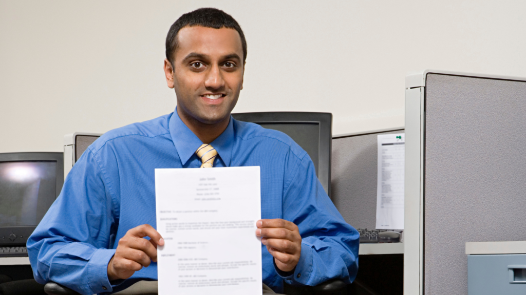 Man of Middle Eastern descent holds up his resume, while sitting in an office cubicle. 