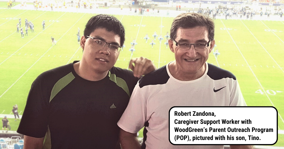 Two men standing in front of a soccer field, one is wearing a black tshirt with green shoulders, the other is wearing a white shirt with black shoulders. Both are wearing glasses.